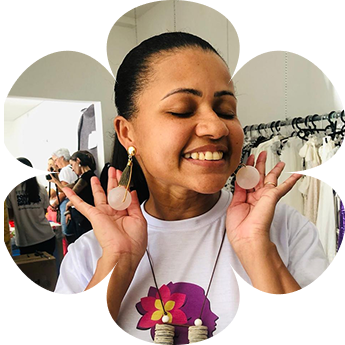 Fabiana Moura, profissional da área da beleza e voluntária do projeto, sorrindo enquanto exibe brincos artesanais, em um ambiente de exposição com roupas ao fundo.